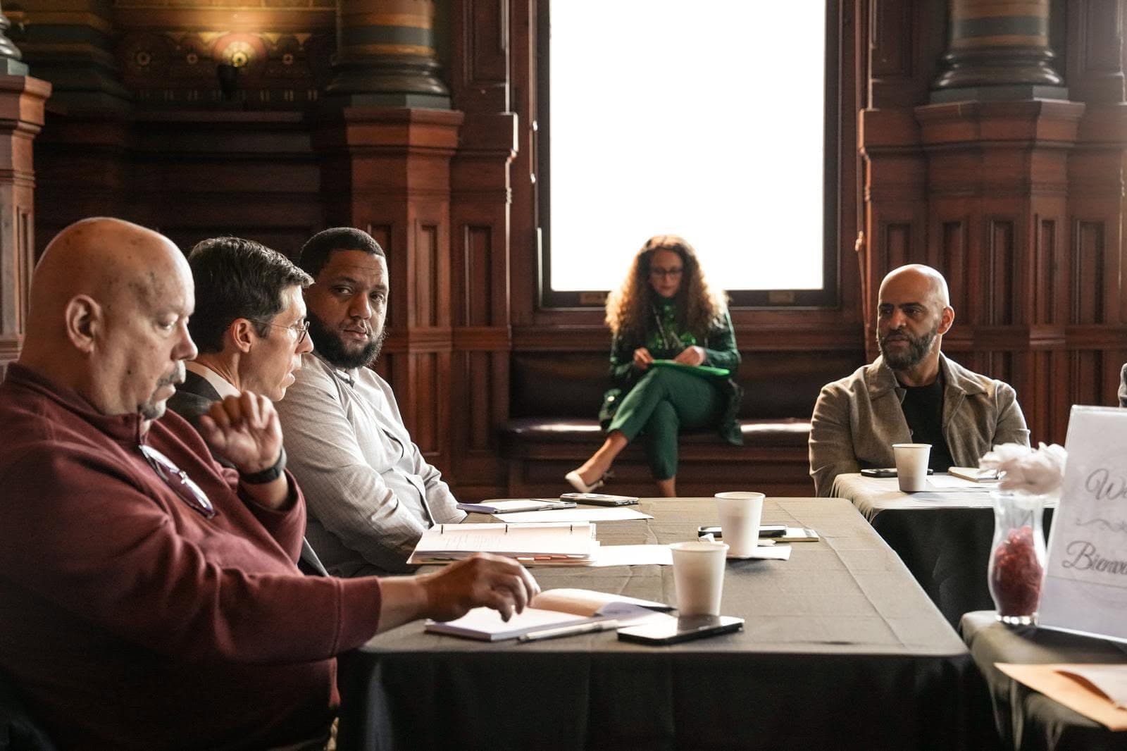 Four people in discussion at a table; a woman sits in the window behind them